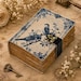 May include: A rectangular journal with a cream-colored cover featuring blue illustrations of two birds perched on branches, surrounded by floral patterns and cursive text. The journal has a black strap with a brass-colored metal latch closure.