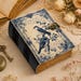 May include: A rectangular journal with a cream-colored cover featuring blue floral borders, two birds perched on branches, and cursive script text. The book has a dark blue spine, a metallic latch closure, and thick, deckled-edge pages.