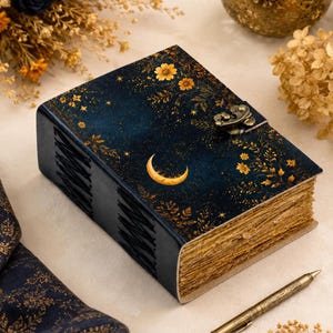 May include: A rectangular journal with a dark blue cover featuring a gold crescent moon, star, and floral pattern. The book has a metallic latch closure, visible side binding with black stitching, and thick, textured, deckled-edge paper pages.