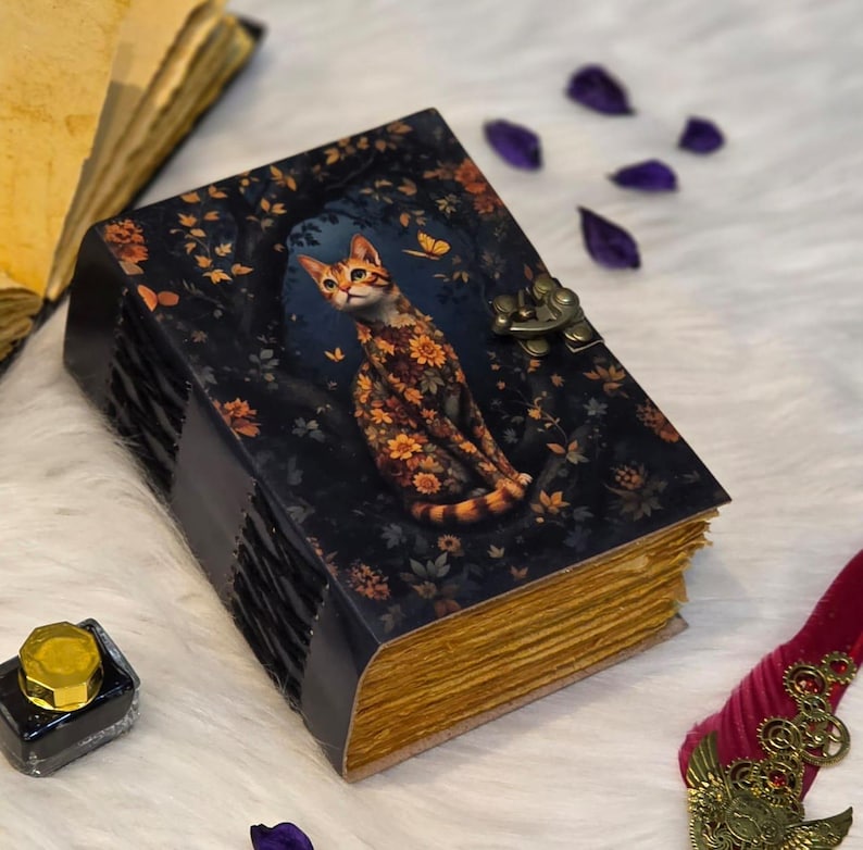 May include: A decorative journal with a cat illustration. The cover is dark blue with a floral design and a cat with a floral pattern. The pages have a gold-colored edge. The journal has a metal clasp and a leather spine.