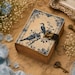 May include: A rectangular journal with a cream-colored cover featuring blue illustrations of two birds perched on branches, floral borders, and cursive script. The journal includes a dark brown leather strap with a brass-colored metal latch closure.