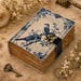 May include: A rectangular journal with a cream-colored cover featuring blue illustrations of two birds perched on branches, surrounded by floral borders and script text. The book has a black strap with a brass-toned metal latch and deckled pages.