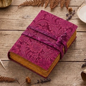 May include: A rectangular journal with a deep magenta cover featuring an embossed floral and leaf pattern. The journal is secured with a matching magenta wrap-around tie. The pages have a weathered, golden-brown appearance with deckled edges.