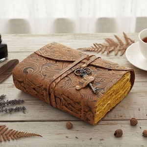 Puede incluir: Un diario de cuero marr&oacute;n con un dise&ntilde;o floral en relieve y un cierre de llave. El diario est&aacute; cerrado con una correa de cuero y una llave decorativa. Las p&aacute;ginas tienen un tono dorado. Una taza de t&eacute; y un tintero est&aacute;n en el fondo.