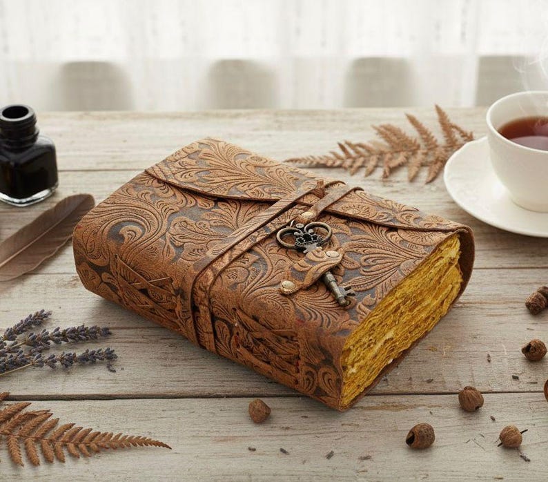 Puede incluir: Un diario de cuero marr&oacute;n con un dise&ntilde;o floral en relieve y un cierre de llave. El diario est&aacute; cerrado con una correa de cuero y una llave decorativa. Las p&aacute;ginas tienen un tono dorado. Una taza de t&eacute; y un tintero est&aacute;n en el fondo.