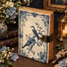 May include: A rectangular journal with a cream-colored cover featuring a blue floral border, two birds perched on a branch, and cursive script text. The journal has a dark brown leather-like strap with a metallic latch and deckled paper edges.