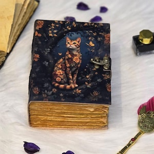 May include: A vintage-style journal with a cat illustration. The cover features a dark blue background with floral patterns and a sitting cat adorned with flower patterns. The journal has a metal clasp and aged-looking pages.