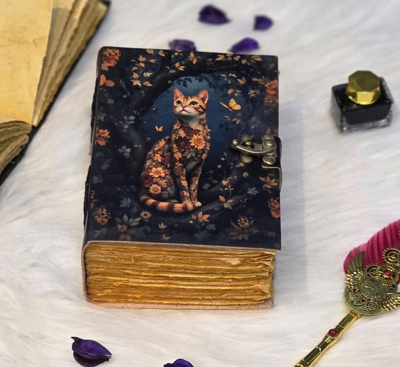 May include: A vintage-style journal with a cat illustration. The cover features a dark blue background with floral patterns and a sitting cat adorned with flower patterns. The journal has a metal clasp and aged-looking pages.