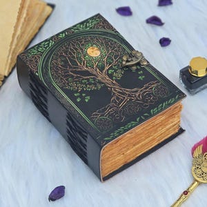 Puede incluir: Un diario de cuero negro con un diseño de árbol de la vida en verde y dorado. El diario tiene un cierre de metal y páginas envejecidas y amarillentas. La portada presenta detalles intrincados y una luna llena.