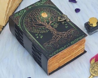 Puede incluir: Un diario de cuero negro con un diseño de árbol de la vida en verde y dorado. El diario tiene un cierre de metal y páginas envejecidas y amarillentas. La portada presenta detalles intrincados y una luna llena.