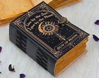 Puede incluir: Un diario de cuero negro con texto e ilustraciones doradas en relieve. La portada dice "Live by the Sun, Love by the Moon" y "Sun of Moon". Las páginas tienen un aspecto vintage envejecido. Un cierre metálico asegura el diario.