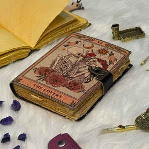May include: A handcrafted journal with a tarot card design. The cover depicts a skeleton couple, flowers, and the words "THE LOVERS." The journal is bound with a black leather strap and has aged pages. Other decorative items are in the background.