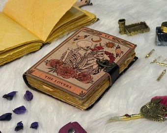 Op de afbeelding: Een handgemaakt notitieboek met een tarotkaartontwerp. De omslag toont een skeletpaar, bloemen en de woorden "THE LOVERS". Het notitieboek is gebonden met een zwarte leren band en heeft verouderde pagina's. Andere decoratieve items staan op de achtergrond.
