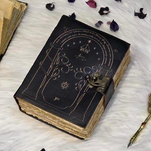 Puede incluir: Diario de cuero negro con un diseño en relieve y cierre de broche. La portada presenta una ilustración con una corona y texto. Las páginas envejecidas tienen bordes dorados. Elementos decorativos: pétalos de flores secas.