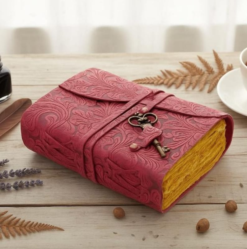 Puede incluir: Un diario de cuero burdeos con un patr&oacute;n floral en relieve. El diario se asegura con una correa y un cierre de llave decorativo. Las p&aacute;ginas tienen un aspecto amarillento y envejecido, lo que sugiere un estilo vintage.