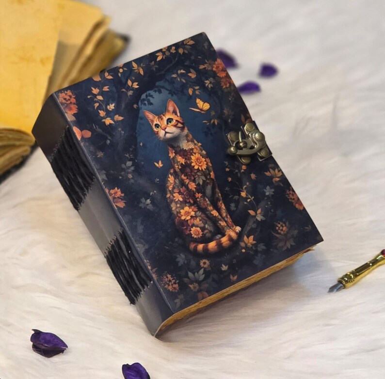 May include: A dark blue journal with a cat illustration. The cat is adorned with floral patterns, set against a backdrop of a forest with a butterfly. The journal has a metal clasp and stitched binding.