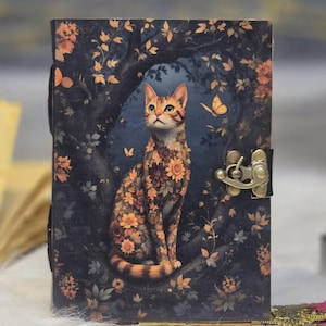 May include: A decorative journal with a cat illustration. The cat is adorned with orange and yellow floral patterns, set against a dark blue background with a forest theme. The journal has a metal clasp and a leather strap.