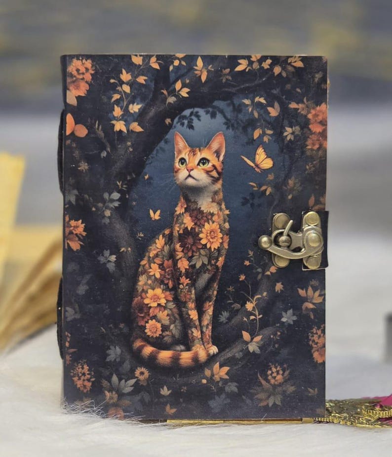 May include: A decorative journal with a cat illustration. The cat is adorned with orange and yellow floral patterns, set against a dark blue background with a forest theme. The journal has a metal clasp and a leather strap.