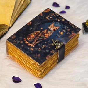 May include: A vintage-style journal with a dark blue cover featuring a cat illustration adorned with floral patterns. The journal has aged, yellowed pages and a metal clasp with a black leather strap. The edges of the pages are also yellowed.