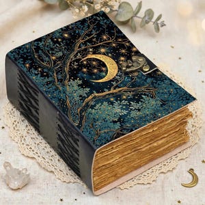May include: A rectangular journal with a dark blue cover featuring a gold crescent moon and star-filled forest illustration. The book has a bronze-colored metal latch closure and thick, deckled-edge pages that appear tan and textured.