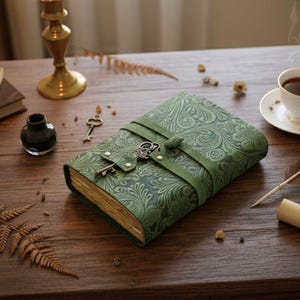 Puede incluir: Diario de cuero verde con un patr&oacute;n floral en relieve y cierre de llave. El diario est&aacute; asegurado con una correa de cuero verde y presenta un dije de llave vintage. Las p&aacute;ginas tienen un tono dorado. Un art&iacute;culo decorativo para escribir o llevar un diario.