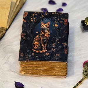 May include: A decorative journal with a cat illustration. The cover features a cat adorned with floral patterns, set against a dark blue background with foliage and a butterfly. The journal has aged, yellowed pages and a clasp closure.
