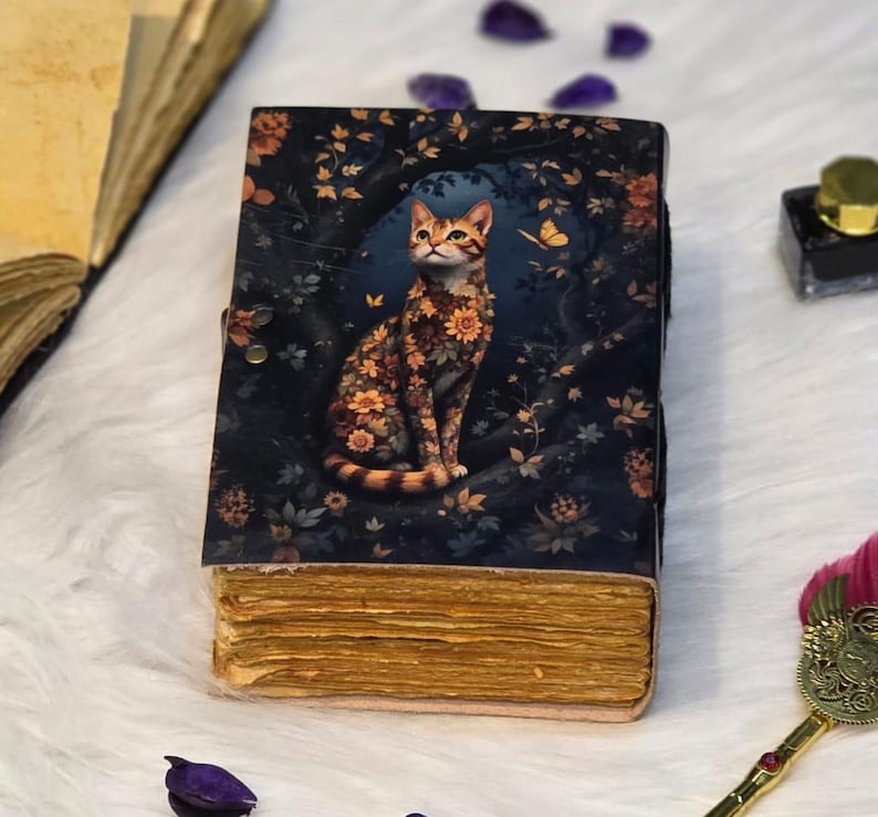 May include: A decorative journal with a cat illustration. The cover features a cat adorned with floral patterns, set against a dark blue background with foliage and a butterfly. The journal has aged, yellowed pages and a clasp closure.