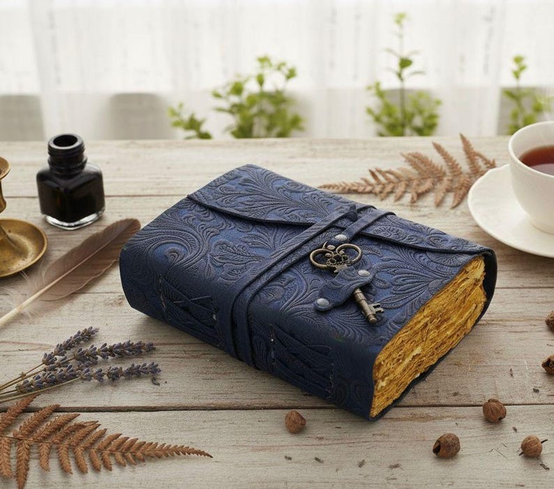 Puede incluir: Un diario de cuero azul oscuro con un patr&oacute;n floral en relieve. El diario se asegura con una correa y un cierre de llave decorativo. Las p&aacute;ginas envejecidas tienen un tono dorado, lo que sugiere una est&eacute;tica vintage.