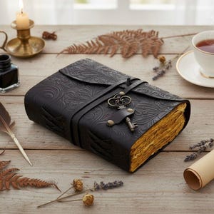 Puede incluir: Un diario de cuero negro con un dise&ntilde;o floral en relieve y un cierre de llave. El diario tiene un cierre de correa y p&aacute;ginas envejecidas. Un dije de llave decorativo cuelga de la correa. El diario est&aacute; sobre una superficie de madera.