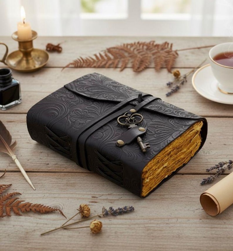 Puede incluir: Un diario de cuero negro con un dise&ntilde;o floral en relieve y un cierre de llave. El diario tiene un cierre de correa y p&aacute;ginas envejecidas. Un dije de llave decorativo cuelga de la correa. El diario est&aacute; sobre una superficie de madera.