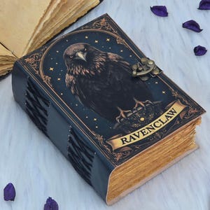 May include: A dark blue leather-bound journal with a detailed illustration of a raven on the cover. The cover also features the word "RAVENCLAW" on a banner. The pages have a gold-colored edge, and the journal is secured with a metal clasp.