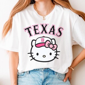 May include: White t-shirt featuring the word "TEXAS" in black and pink lettering above a cartoon cat wearing a baseball cap with a pink bow. The cat has black whiskers and a black outline.