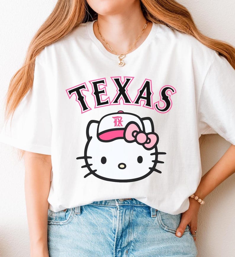 May include: White t-shirt featuring the word "TEXAS" in black and pink lettering above a cartoon cat wearing a baseball cap with a pink bow. The cat has black whiskers and a black outline.