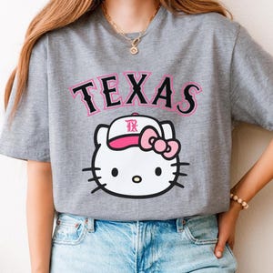 May include: Gray t-shirt featuring the word "TEXAS" in pink and black lettering above a white cartoon cat wearing a baseball cap with a pink bow. The cat has black whiskers and eyes.