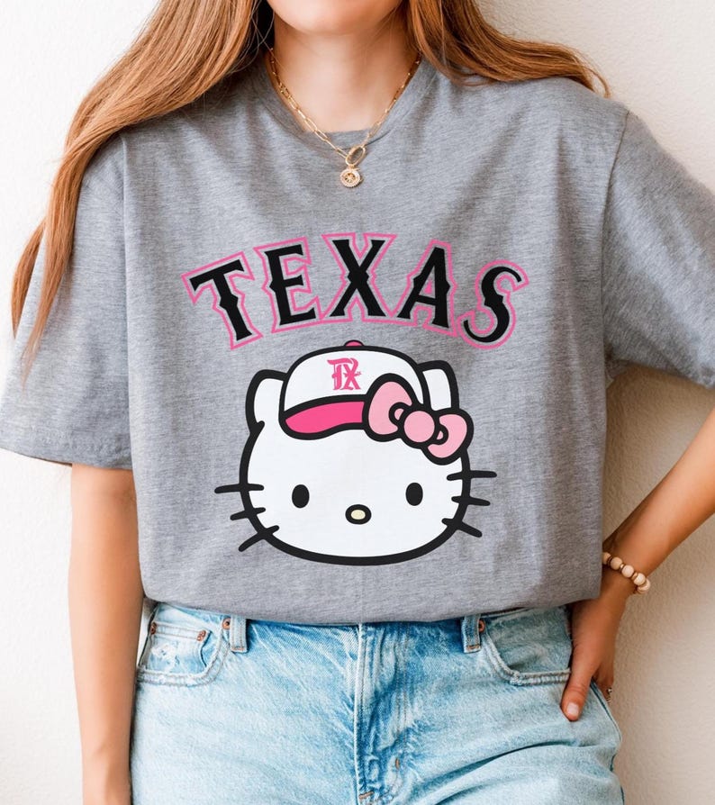 May include: Gray t-shirt featuring the word "TEXAS" in pink and black lettering above a white cartoon cat wearing a baseball cap with a pink bow. The cat has black whiskers and eyes.