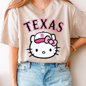 May include: Beige t-shirt featuring the word "TEXAS" in black and pink lettering above a white cartoon cat wearing a pink bow and a baseball cap with the letters "TX". The shirt is paired with blue jeans.