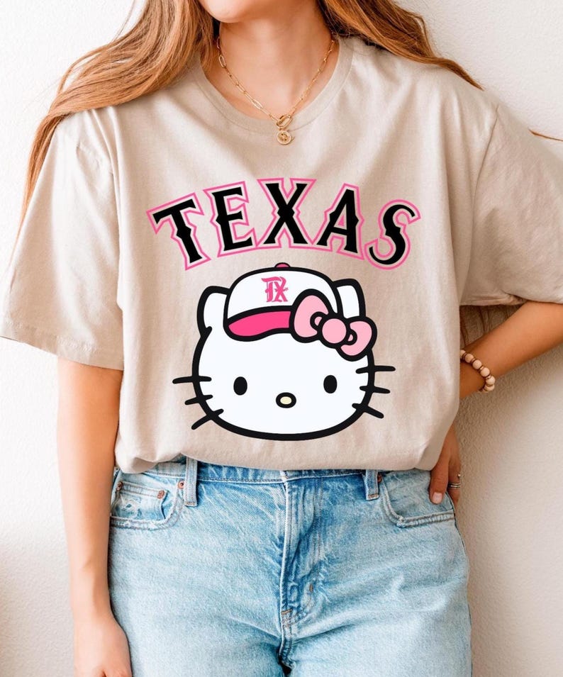 May include: Beige t-shirt featuring the word "TEXAS" in black and pink lettering above a white cartoon cat wearing a pink bow and a baseball cap with the letters "TX". The shirt is paired with blue jeans.