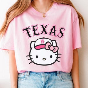 May include: Pink t-shirt featuring the word "TEXAS" in black and pink lettering above a white cartoon cat face wearing a pink hat and bow. The cat has black whiskers and black eyes. The shirt is a light pink color.
