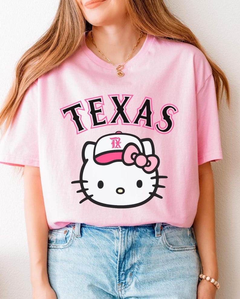 May include: Pink t-shirt featuring the word "TEXAS" in black and pink lettering above a white cartoon cat face wearing a pink hat and bow. The cat has black whiskers and black eyes. The shirt is a light pink color.