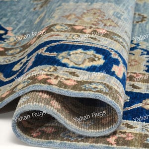 May include: A rolled-up blue rug with a detailed pattern. The rug features a border with a dark blue design and floral accents in pink and beige. The text "Yafiah Rugs" is repeated across the rug's surface.