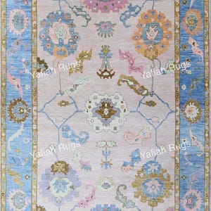 May include: A rectangular rug with a light pink central field and a blue border. The rug features floral and geometric patterns in shades of blue, orange, pink, and green. The text "Yafiah Rugs" is repeated along the border.
