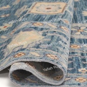 May include: A rolled-up blue rug with a detailed pattern. The rug features a mix of light and dark blue hues, with beige and brown floral and geometric designs. The rug's edge is a lighter blue, and the brand name "Yafiah Rugs" is printed on the rug.