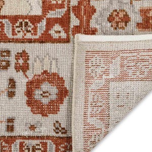 May include: Close-up of a beige and rust-colored rug with a detailed geometric pattern. The rug features floral and rectangular designs in shades of rust and beige. The corner of the rug is folded over, revealing the edge with a border.