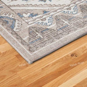 May include: Close-up of a corner of a patterned rug with a beige, blue, and white design. The rug has a border and is placed on a wooden floor. The text "Yafiah Rugs" is visible on the floor.