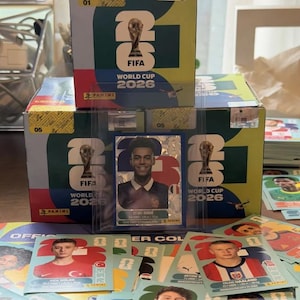 Könnte beinhalten: Ein Panini FIFA World Cup 2026 Sammelkarten-Boxset mit einer Desiré Doué-Karte in einer Schutzhülle. Die Box zeigt das FIFA World Cup 2026 Logo und Branding. Mehrere lose Fußball-Sammelkarten sind im Vordergrund verstreut zu sehen.