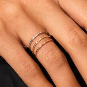 Könnte beinhalten: Ein Set aus drei dünnen, glatten Stapelringen an einem Finger. Die Ringe haben drei verschiedene Metalltöne, darunter ein silberfarbenes Band, ein gelbgoldfarbenes Band und ein roségoldfarbenes Band, alle mit einer polierten Oberfläche.