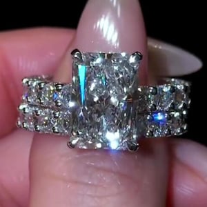 Może przedstawiać: Pierścionek zaręczynowy z diamentem o szlifie radiant, z dużym prostokątnym kamieniem centralnym osadzonym w srebrzystej metalowej obrączce. Obrączka jest wysadzana mniejszymi diamentami o szlifie okrągłym na całej widocznej części szyny.