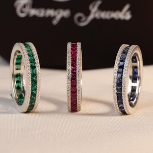 May include: Three silver eternity bands featuring channel-set square-cut gemstones in green, red, and blue, flanked by rows of small clear stones. The rings are positioned in front of a black box with the white text Orange Jewels printed on it.