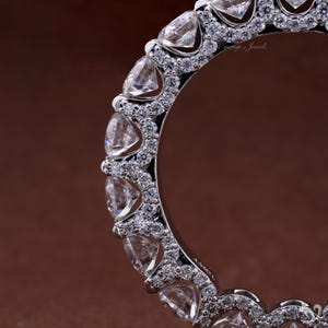 Puede incluir: Primer plano de un anillo de plata con una fila de piedras preciosas claras de corte triangular. Piedras preciosas redondas más pequeñas están engastadas entre las piedras más grandes. El anillo está sobre un fondo marrón. El texto "Orange Jewels" es visible.