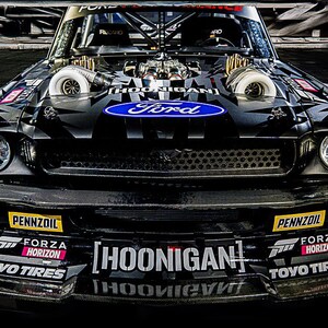Puede incluir: Coche de carreras Ford Mustang Hoonigan negro y gris con un logotipo azul de Ford. El coche tiene dos grandes turbocompresores, un alerón delantero con logotipos de patrocinadores y el logotipo de Hoonigan. Los faros tienen un diseño en forma de X.